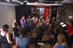 A small rehearsal studio with a big group of people standing together. A group of 5 presenters face the rest of the crowd, discussing in ASL to a watchful crowd.