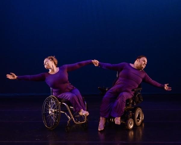 On stage, Frank Hull in his powerchair holds his arms extended and connected with dance partner in a manual wheelchair.