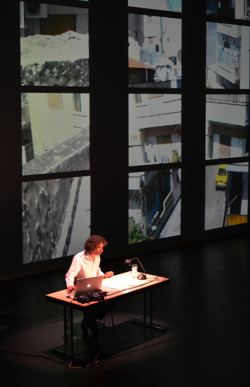 Rabih Mroué sitting at a desk on stage, with several square projections of video behind him.