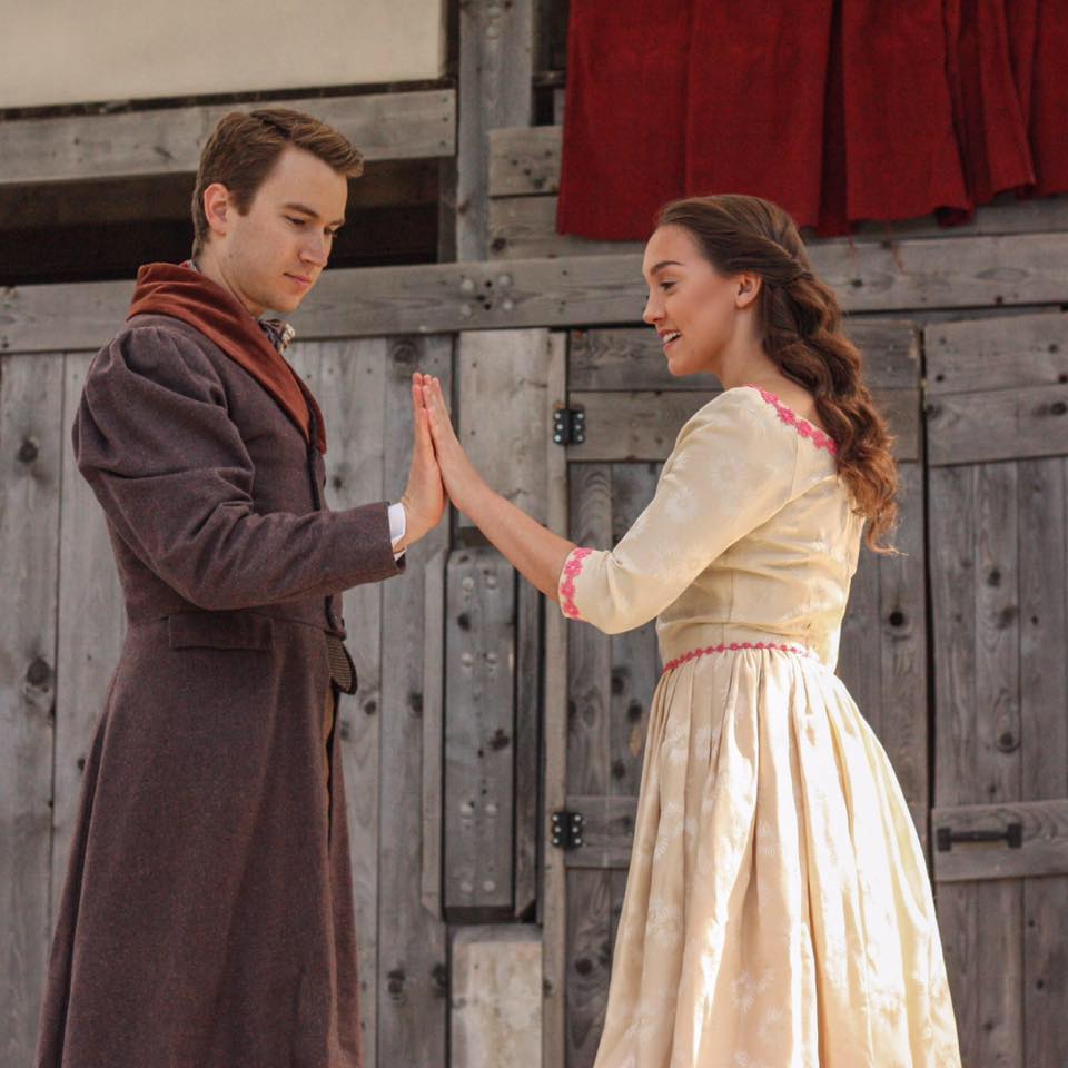 Man and woman standing on a wooden stage, with one hand reached towards the other's. Palms touching.