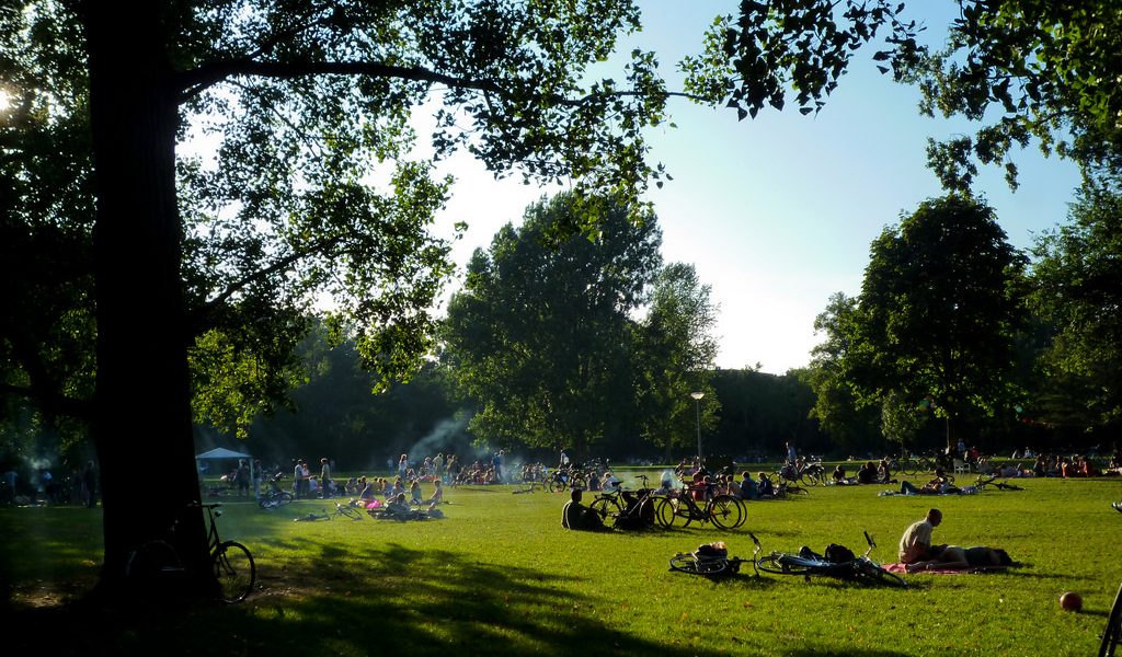 Lush city park in the summer time. People lounging in grass, crossing on bikes, bbqing.