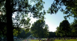 Lush city park in the summer time. People lounging in grass, crossing on bikes, bbqing.