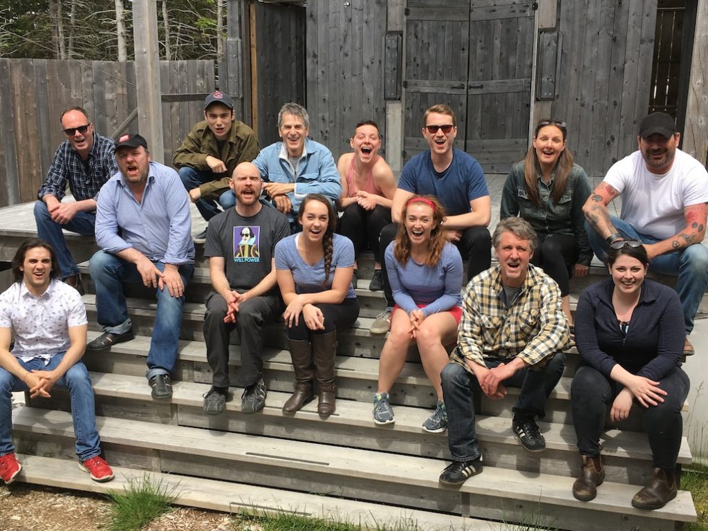 Goofy shot of large cast sitting infront of wood cabin.