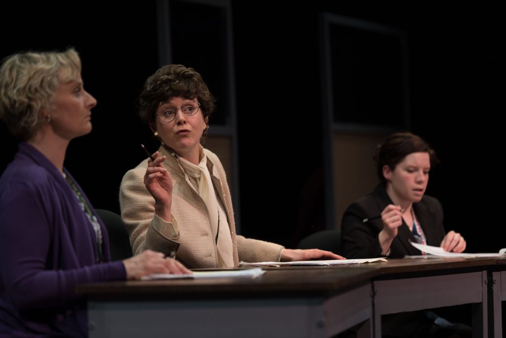 3 women dressed in 60s clothing discuss at desks.