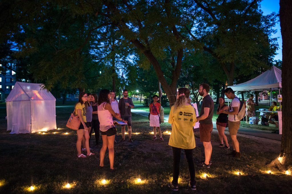 Group of people standing in a circle between colourful tents and trees.