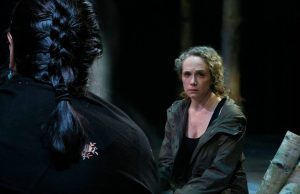 Peering over the shoulder of a man with braided hair. He faces a dejected looking woman with curly blonde hair.
