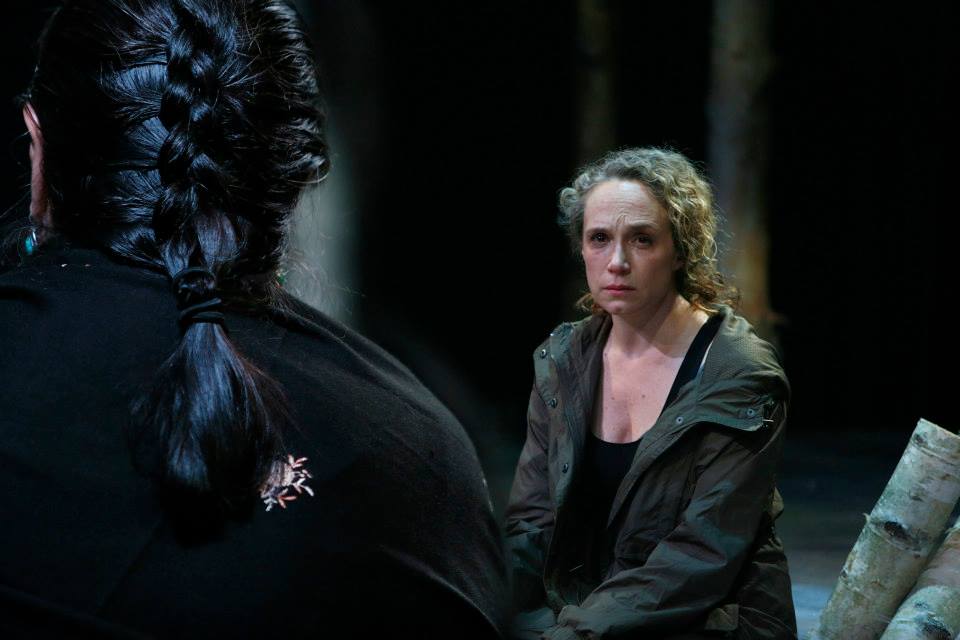 Peering over the shoulder of a woman with braided hair. He faces a dejected looking woman with curly blonde hair.