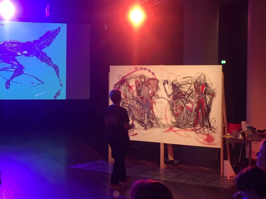 Colourful light stage with an artist painting a large canvas. Their painting is references images of wheelchairs with expressive lines.