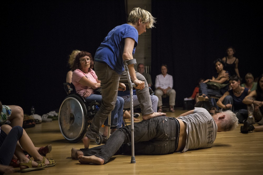Audience seated casually around performers in centre of floor. One performer using crutches steps onto the hip of another performer laying on the floor.