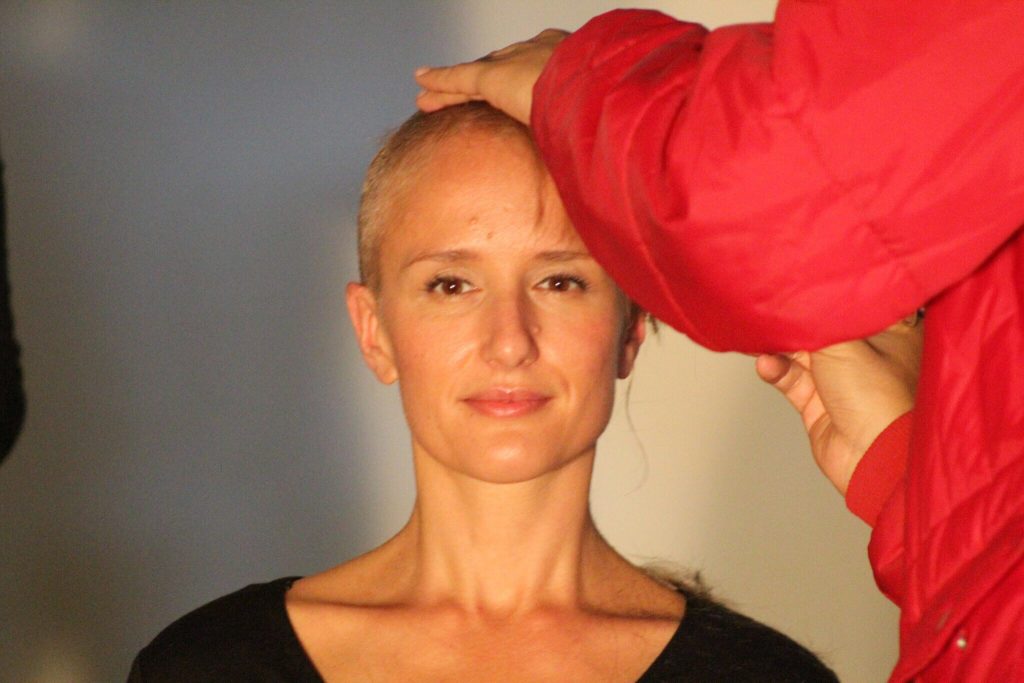 Clare staring right into camera with a freshly shaved head. Barber adds finishing touches with razor.