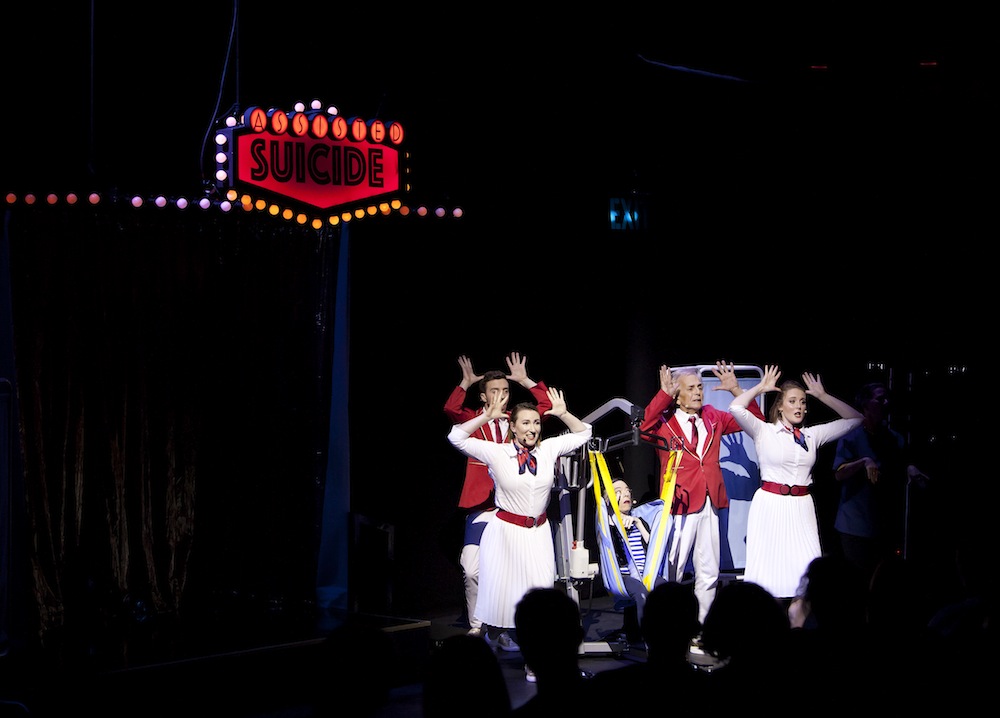 Production still of 5 performers dressed in 50s style clothing, singing with their hands posed by their foreheads. A sign reads 'Suicide" behind them.