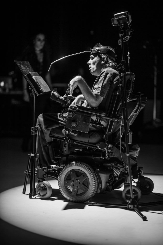 Man in a wheelchair on stage, performing with a live feed camera and letter board.