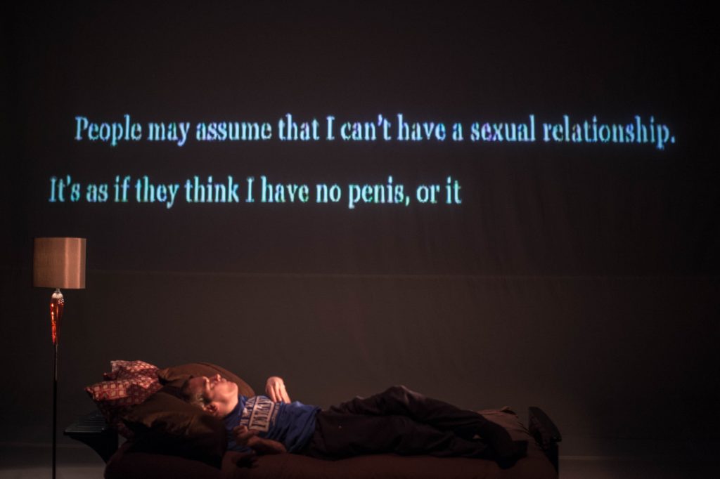 A man laying on a long couch, with words projected above him on the wall behind. Words read: People may assume that I can't have a sexual relationship. It's as if they think I have no penis, or it. 