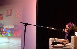 Elaine sits on a comfy white chair on stage, speaking into a microphone. Behind her is a projection screen with an image of a couch on it.