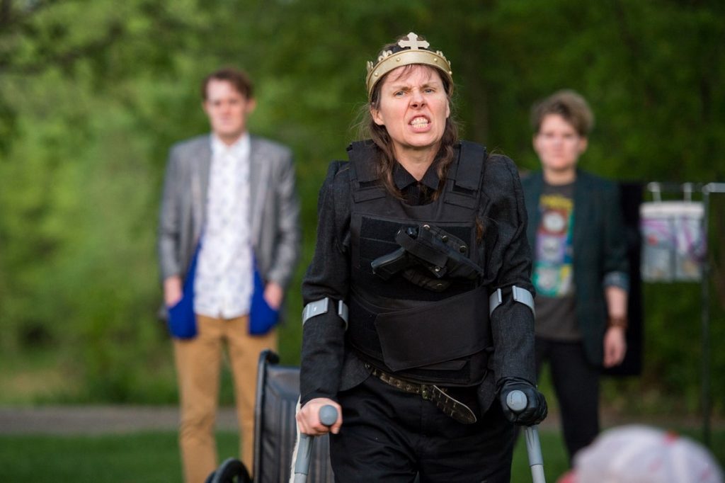 3 actors perform outdoors, under sunny green trees. Debbie is centre, snarling at the audience in a golden crown. She is using silver crutches.