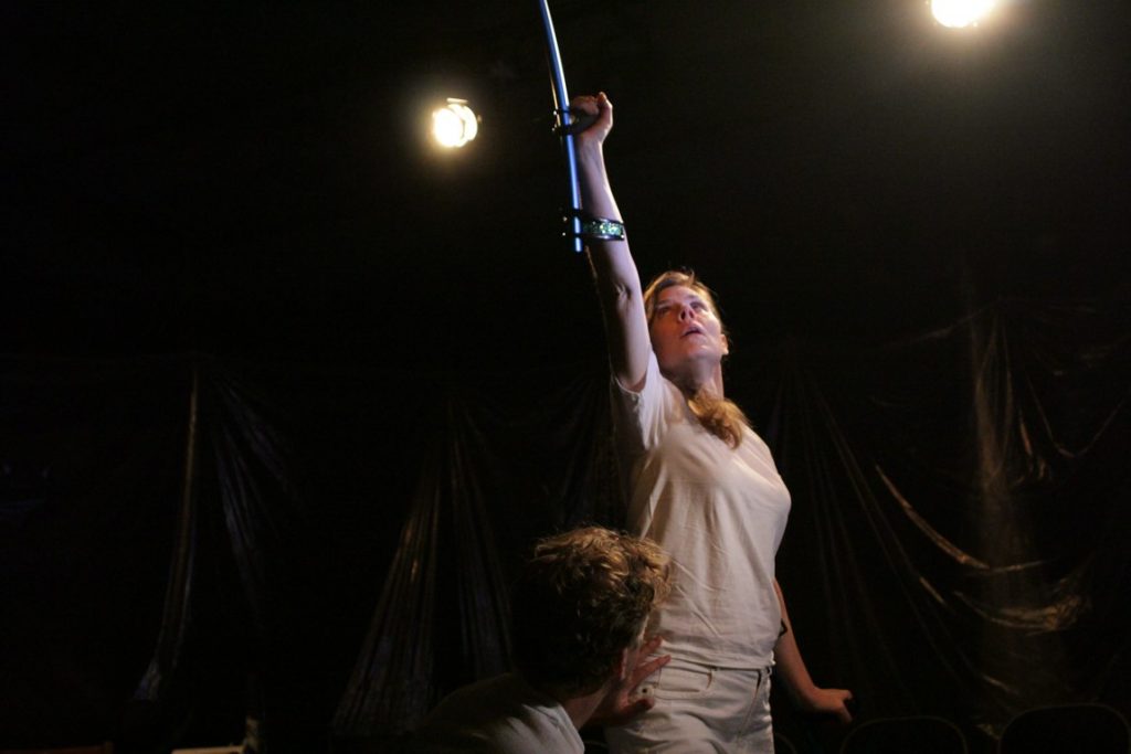 Debbie stands on stage in all white, with one arm extended above her pointing her crutch up to ceiling.