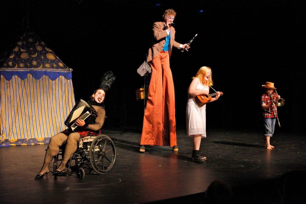 4 performers on stage in a circus style show. One on stilts, Debbie using a wheelchair, all playing instruments.