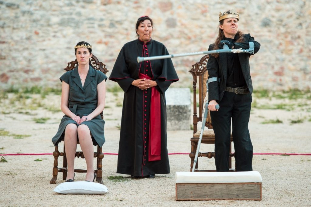 3 actors perform in a sandy outdoor space, in front of a stone wall. Debbie, in a crown, angrily points towards another crowned actor who is seated. An actor dressed like a bishops laughs in the background.