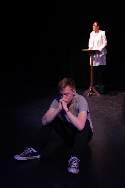 Downstage, Jonathan sits on the ground with his hands folded in prayer in front of his mouth. Upstage, a woman in all white stands at a pulpit.