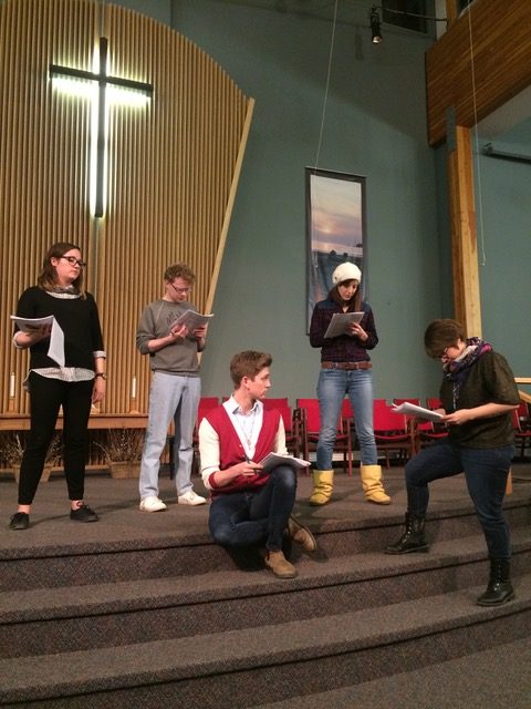 4 actors and a dramaturg gather at the front of a church with scripts in hand. An illuminated cross hangs above their heads.