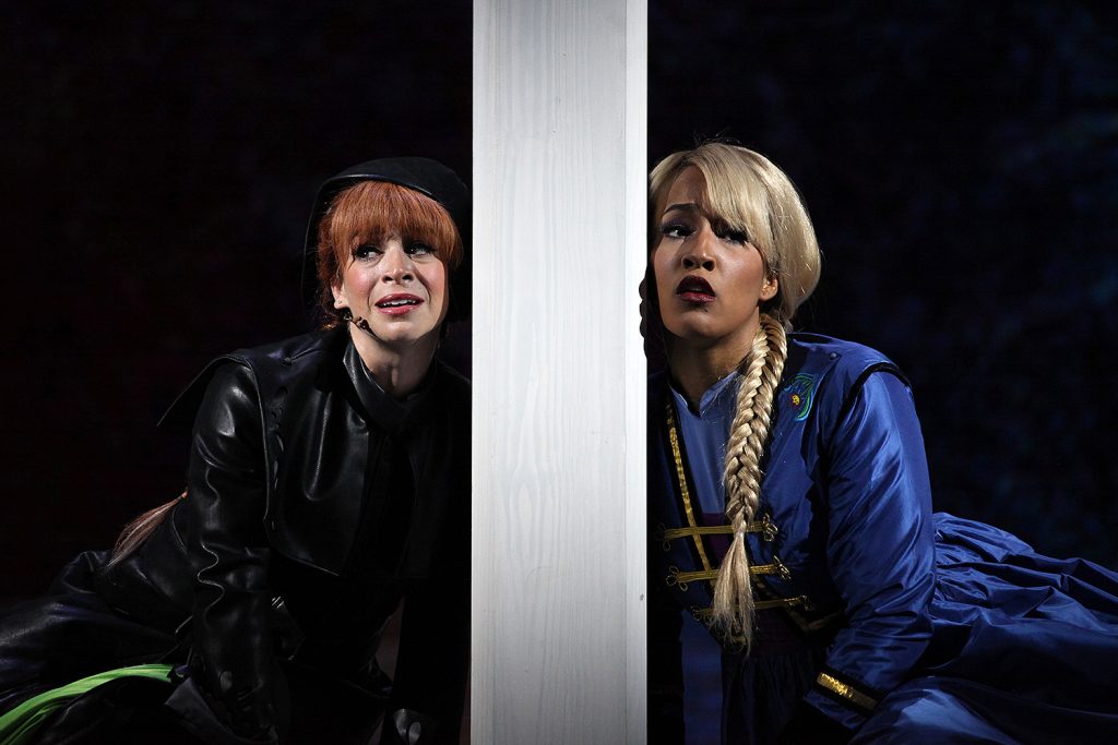 From a stage production of Disney's Frozen Live. Two actresses singing, facing the audience, and leaning on a large post between them.