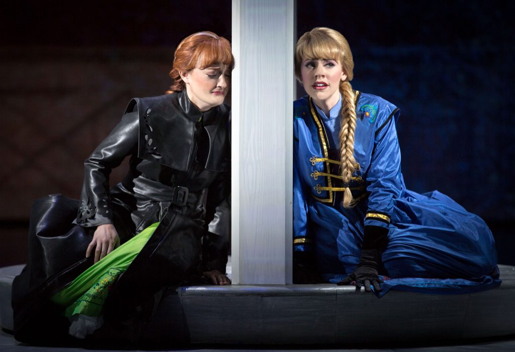 From a stage production of Disney's Frozen Live. Two actresses singing, facing the audience, and leaning on a large post between them.