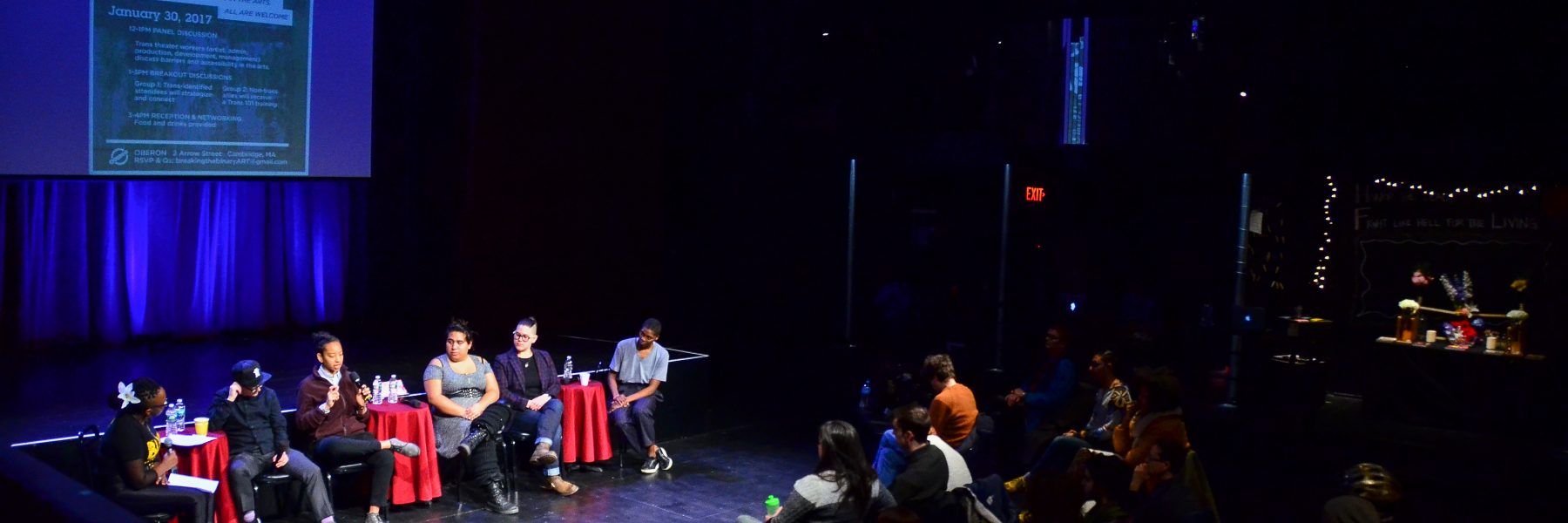 A panel of 6 speakers sits in front of a lit stage. An audience watches them attentively in the darkness.
