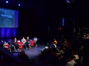 A panel of 6 speakers sits in front of a lit stage. An audience watches them attentively in the darkness.