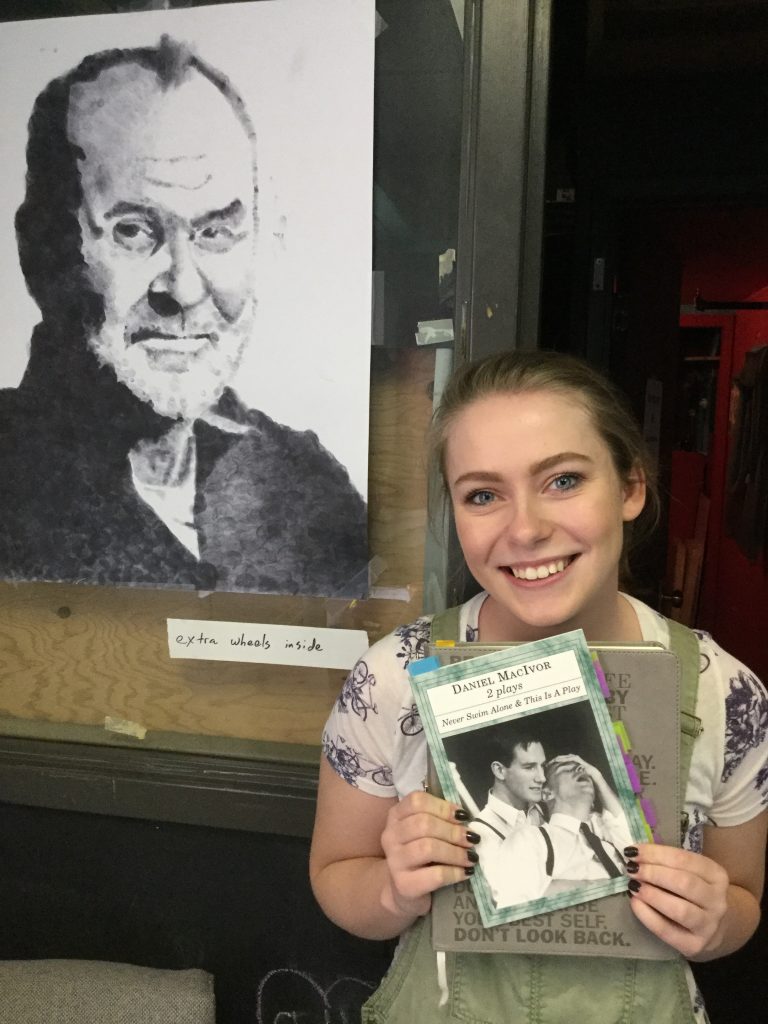 Jane, high school girl, holds a copy of Never Swim Alone by Daniel MacIvor while standing in front of a portrait of the author. 