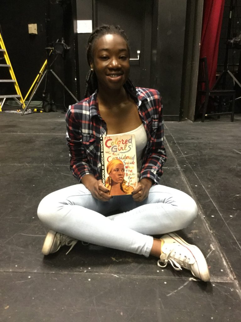 Joy, high school girl, holding a copy of the script For Coloured Girls Who Have Considered Suicide When The End of the Rainbow is not Enough