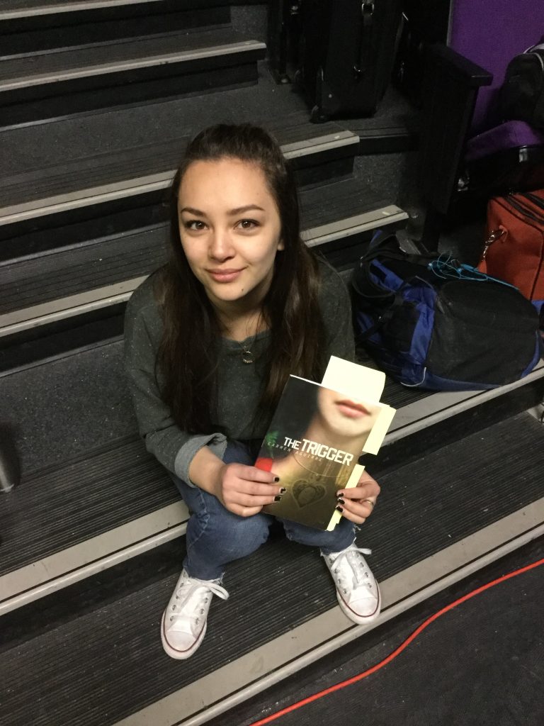 Haylee, a high school girl, is holding a copy of the script for The Trigger by Carmen Aguirre.