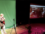 Sarah Garton Stanley stands in front of a green screen, video monitor shows people gathered in the digital rehearsal hall.