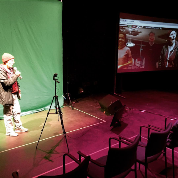 Sarah Garton Stanley stands in front of a green screen, video monitor shows people gathered in the digital rehearsal hall.