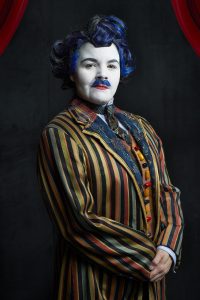 A young woman, her face painted white, dressed in a colourfully striped men's suit with blue hair and a blue mustache. 