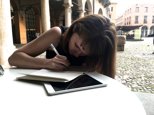 A woman sits in a plaza, leaning over a journal and ipad as she writes. 