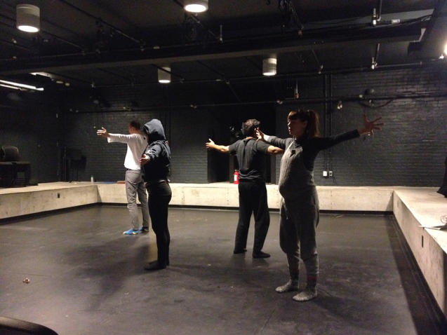 Four actors stand in a cluster in the middle of a blackbox theatre, with their arms extended wide. 