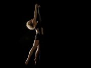 A woman with curly blonde hair arches her back as she holds herself in mid-air by two long fabric silks, against a black background.