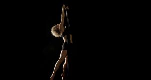 A woman with curly blonde hair arches her back as she holds herself in mid-air by two long fabric silks, against a black background.