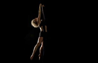 When Circus Meets Theatre: A Tale of Two Lovers A woman with curly blonde hair arches her back as she holds herself in mid-air by two long fabric silks, against a black background.