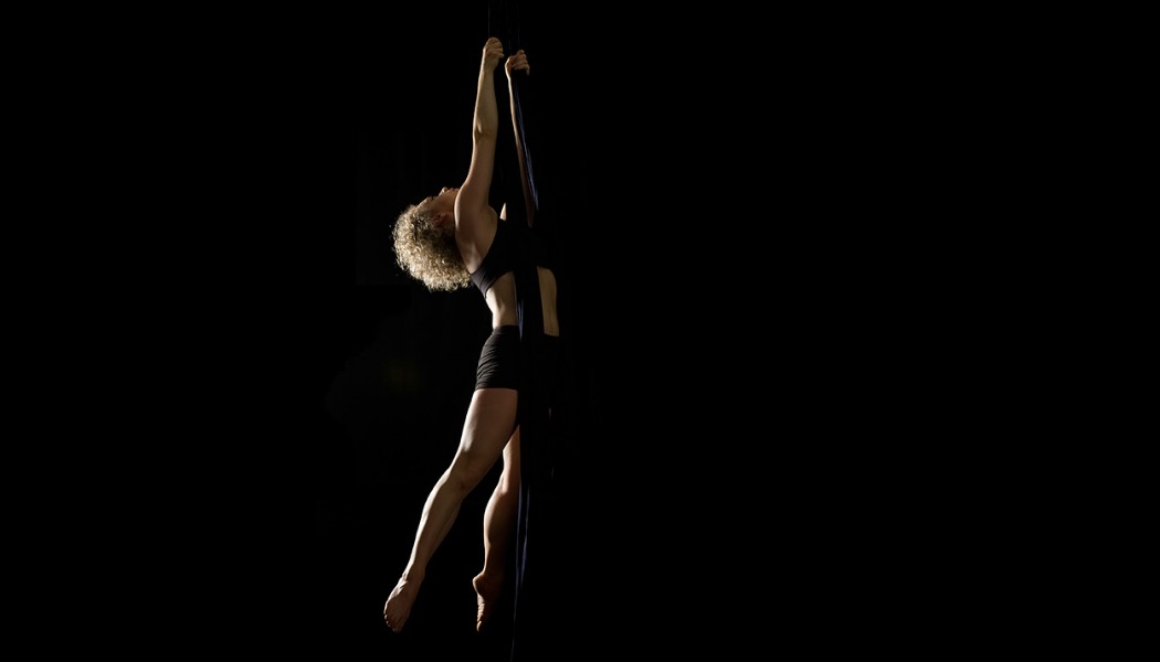 A woman with curly blonde hair arches her back as she holds herself in mid-air by two long fabric silks, against a black background. 