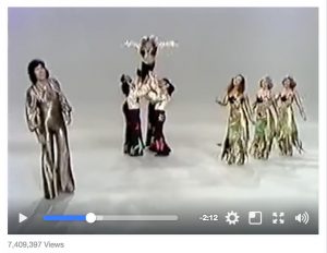 Screen capture of a vintage Spanish music video. One man in gold lameé jumpsuit sings, four men link arms to throw a third into the air with his arms outstretched like a bird, three women in sparkly gowns dance in unison.
