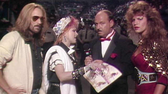 Four figures gathered around a microphone. Cyndi Lauper speaks into the microphone as the other three individuals look at the camera, somewhat surprised.